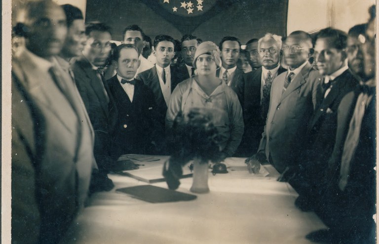 Alzira Soriano - primeira prefeita da América Latina Foto sépia de grupo de pessoas atrás de mesa com vaso de flores. Ao fundo, bandeira do Brasil an...