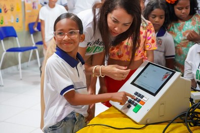 A imagem mostra uma criança sorridente, usando óculos e uma camisa branca com detalhes em azul, ...