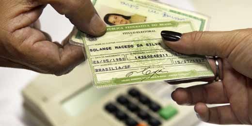 Titulo eleitoral Eleitora passando título eleitoral e RG para mésário com leitor biométrico ao fundo.